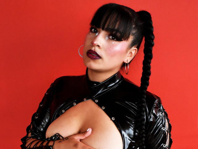 KiaraThomsomp con una blusa de látex negra, sosteniendo sus pechos, fondo rojo, cabello negro trenzado, lápiz labial púrpura, aros grandes, expresión seria, tatuaje en la mano derecha..