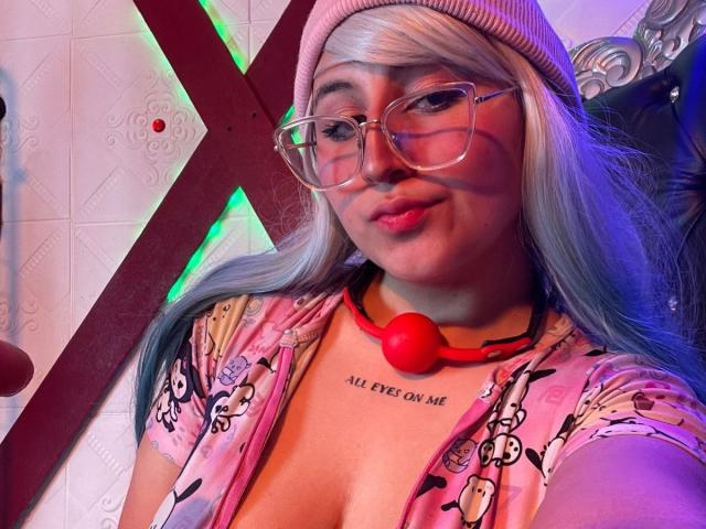 MaddieLesYoux with glasses, pink beanie, and pink hair, wearing a floral top, red ball gag, and "ALL EYES ON ME" tattoo, smiling with pouty lips.