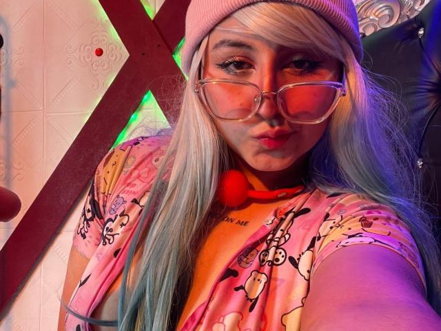 MaddieLesYoux with pink beanie, clear glasses, and pink floral top, showing pouty lips, long blue-and-white hair, and a red ball necklace, against a colorful, cross-shaped background.
