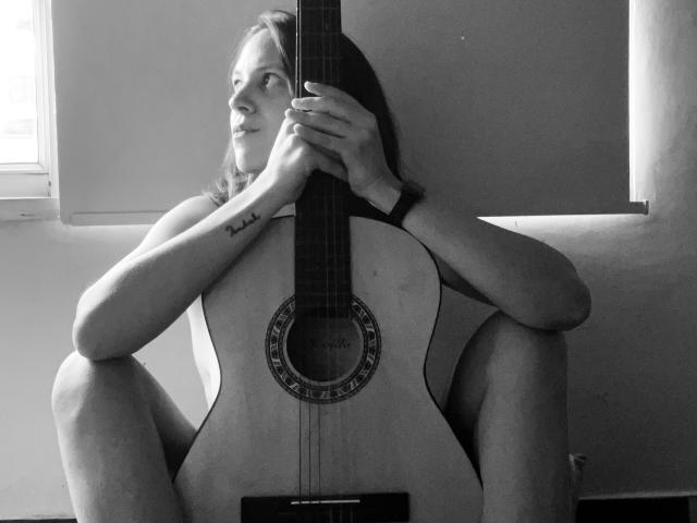 LunaFiorella con una guitarra, sentada con las rodillas levantadas, sosteniendo el mástil de la guitarra acústica, con un tatuaje en el brazo izquierdo, un reloj en la mano derecha, en blanco y negro, mirando hacia un lado, plano medio..