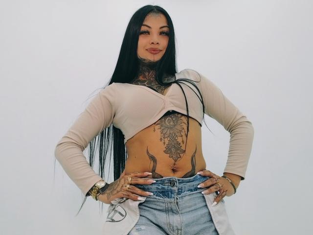 LisaMont with long black hair, tan crop top, and blue jeans, showing off tattoos, including a large chest piece and belly art, hands on hips, smiling confidently.