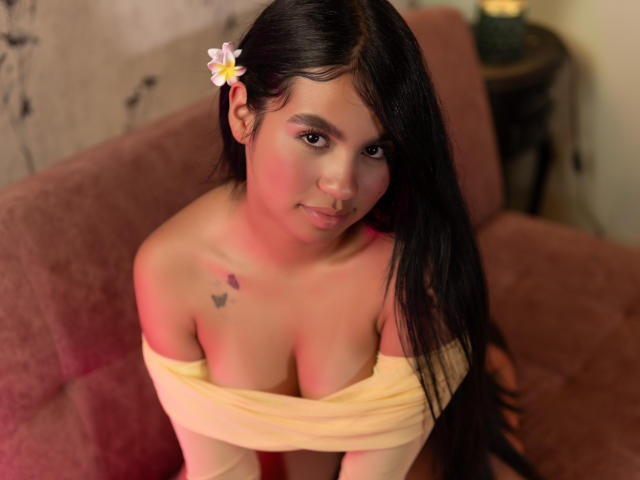 DalilaTennesse with long black hair, a pink flower in her hair, off-shoulder yellow top, showing her tits, tattoo on left shoulder, pinkish-red light, sitting on a pink couch.