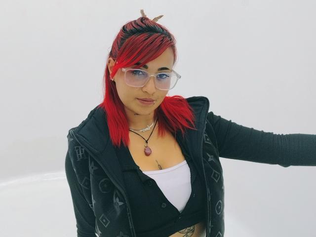 Sushy mit roten Haaren, Brille, schwarzer Jacke, weißem Oberteil, kleinen Brüsten, einer Halskette, die einen Arm ausstreckt, lächelnd, vor weißem Hintergrund, mit dezentem Make-up..