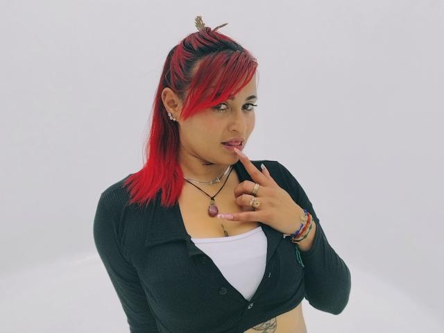 Sushy, com cabelo ruivo, cardigã preto, blusa branca, dedo na boca, mostrando os seios, colar, anéis, pulseiras, tatuagens e uma expressão adorável. Ela é linda e sexy..