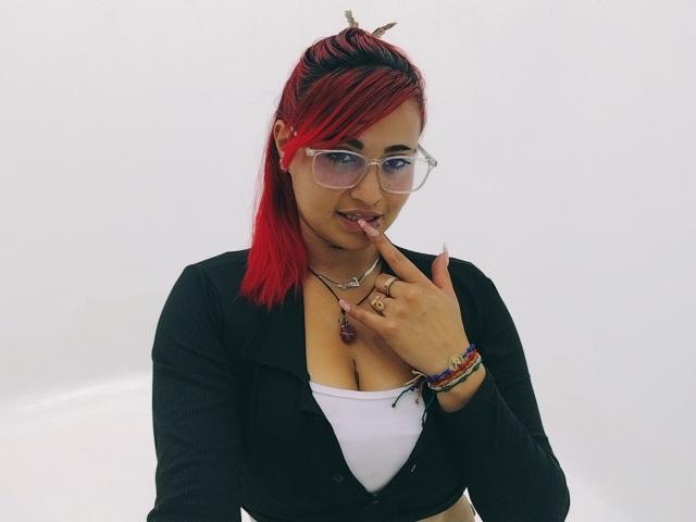 Sushy with red hair, glasses, and black cardigan, finger in mouth, showing tits in white top, bracelets, and rings.