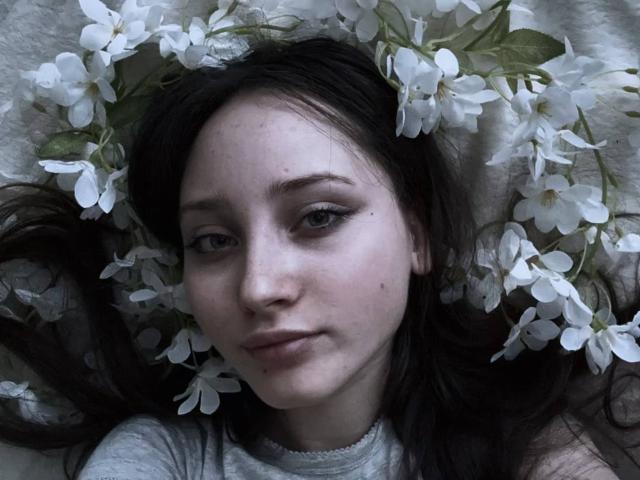 HaileyMuskopf's face, with white flowers in her dark hair, has a slight smile and subtle makeup, wearing a grey top, lying down with a soft, white textured background.
