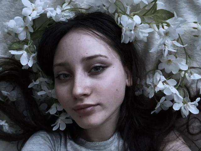 Il viso di Hailey Muskopf, con fiori bianchi tra i capelli scuri, indossa una camicia grigia, appare leggermente triste con un trucco delicato e un sorriso dolce. La sua pelle è pallida..