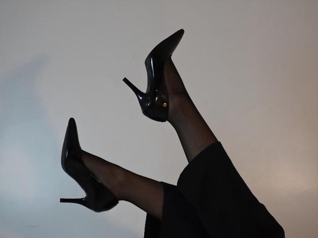 SpencerLe trägt schwarze High Heels mit spitzer Spitze, glänzendem Finish und Stilettoabsätzen, zusammen mit transparenten schwarzen Strumpfhosen, vor einem schlichten Hintergrund..
