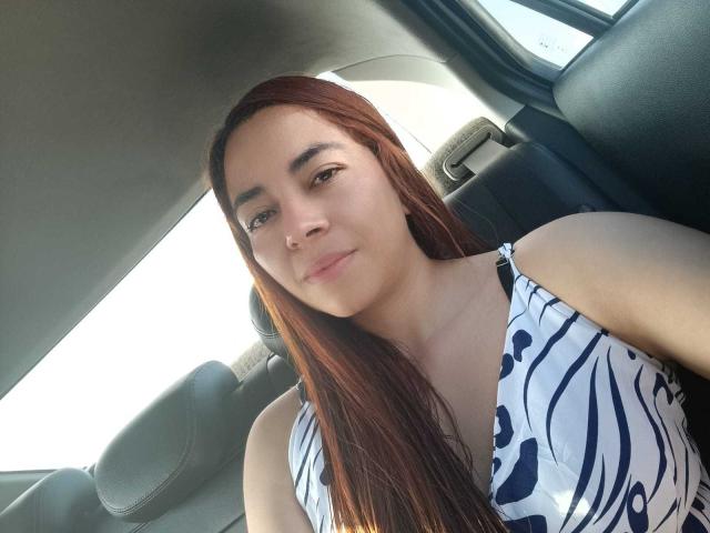 MadisonKoper with long red hair, wearing a white and blue zebra-patterned dress, sitting in a car, showing a slight smile, and looking at the camera.