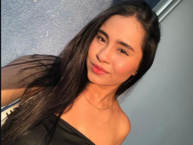 Hanna Harper a de longs cheveux noirs, une peau brun clair et de petites seins. Elle porte un haut noir à une épaule, avec un léger sourire et un fin collier en or..
