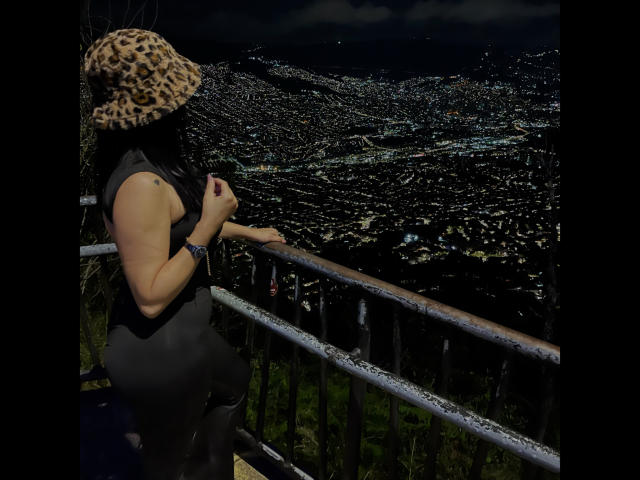 MiaAylen, portant un chapeau léopard, un haut noir sans manches et un pantalon noir, se tient sur une rambarde en métal, regardant une vue nocturne illuminée de la ville, ses fesses légèrement relevées, un ciel sombre au-dessus..