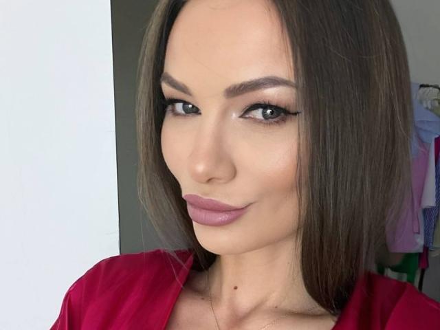 AliceMarr has brown hair, green eyes, and pink lipstick, wearing a red shirt. She's looking at the camera with a slight smile, showing a small beauty mark.