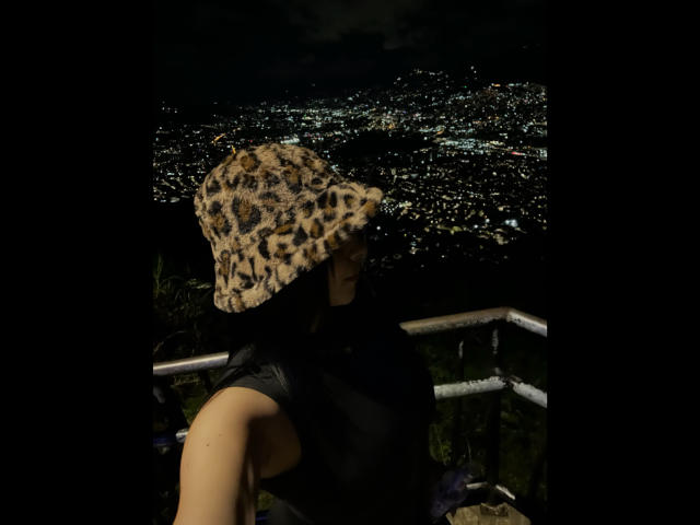 Mia Aylen mit dunklem Haar, die einen Eimerhut mit Leopardenmuster, ein schwarzes Top ohne Ärmel trägt und neben einem Metallgeländer steht, blickt auf eine Stadt voller Lichter in der Nacht..
