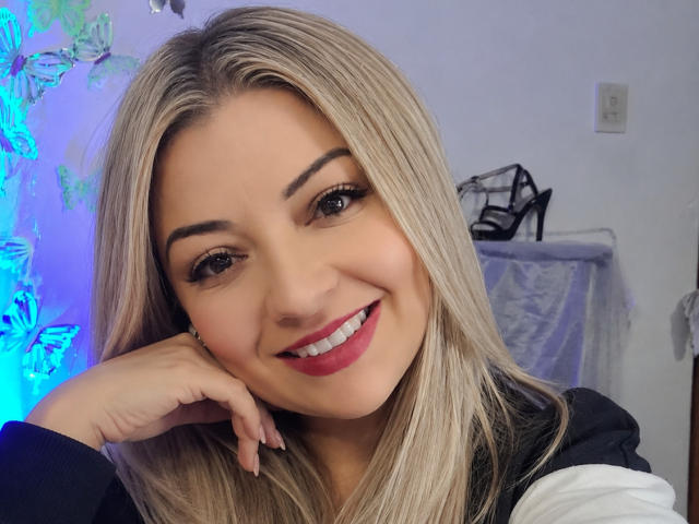 NatalyHami with blonde hair, smiling, red lipstick, black eyebrows, white shirt, hand on cheek, black high heels in background, blue butterfly lights, light skin, cute, attractive.
