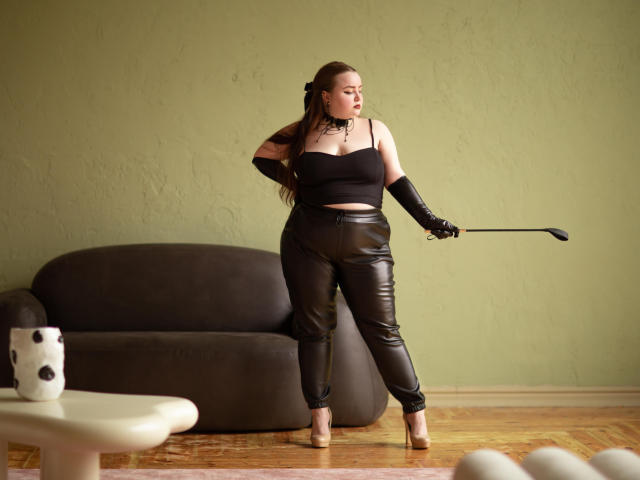 MissEmmar with brown hair, black crop top, leather pants, long gloves, holding a whip, standing in front of a dark couch, wooden floor, and green wall.