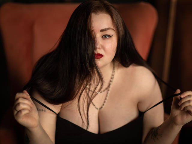 MissEmmar with long black hair, red lipstick, and black dress, pulling straps, showing big tits, chain necklace, and tattoos, sitting in a red chair, looking at camera.