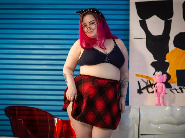 MissEmmar with pink hair, glasses, black bra, red plaid skirt, showing her ass, tattoos, and black cat ear headband, standing in front of blue and white background.