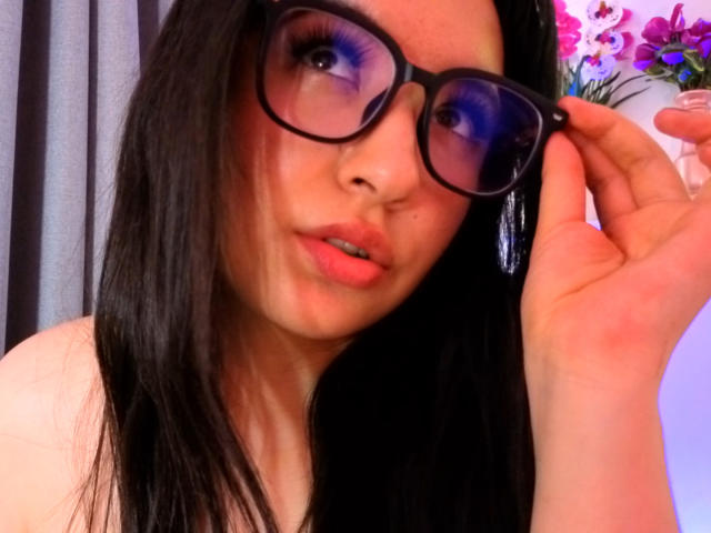 SusanMonroy with black hair, purple-tinted glasses, and pink lips, holding her glasses, showing her face and upper chest, wearing a white top, in a colorful background.
