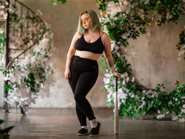 MissEmmar with green hair, black sports bra, and high-waisted black pants, holding a metal pole, standing with a confident stance, wearing black shoes, and a tattoo on her arm.