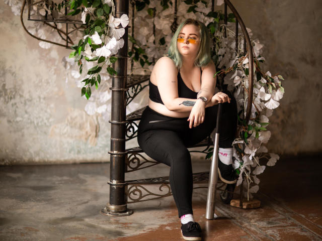 MissEmmar with green hair, yellow glasses, black sports bra, and pants, sitting on a spiral metal bench with white flowers, black shoes, and white socks.