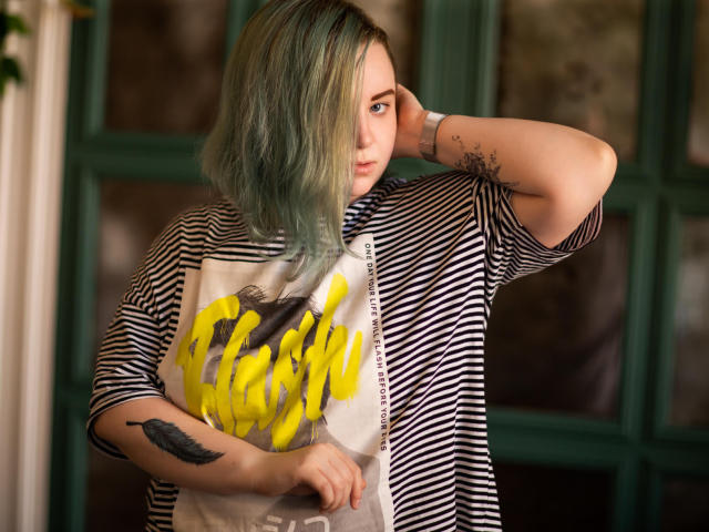 MissEmmar with greenish-blue hair, striped shirt, and yellow graffiti graphic, hand on head, tattoo on forearm, looking serious, standing in front of green window.