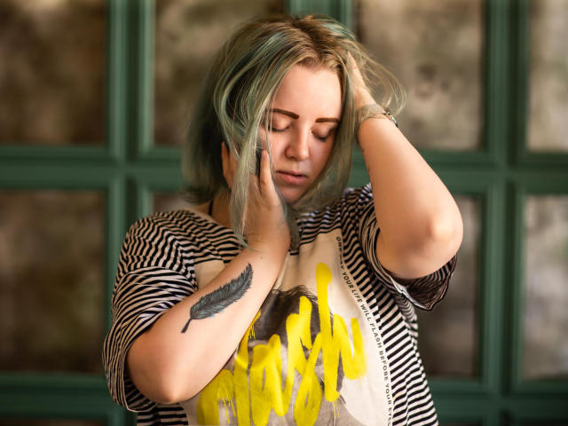 MissEmmar with greenish-blonde hair, hands in her hair, looking down, wearing a striped shirt with yellow paint splatter, feather tattoo on her right wrist.