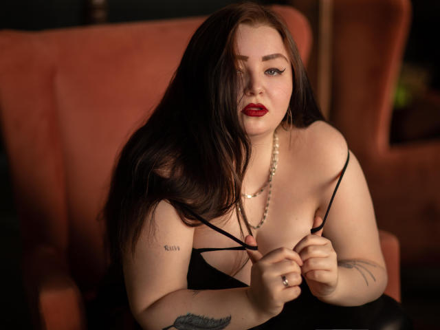 MissEmmar with long brown hair, red lipstick, and pearl necklace, pulling down black straps to expose her tits, sitting in an orange chair, showing tattoos on her arm.