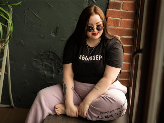 MissEmmar with pale skin, red lipstick, black t-shirt, pink pants, tattoos on arms, sitting on stool, brown hair, sunglasses, brick wall, green textured wall, and potted plant.