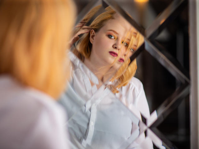 Blonde MissEmmar with fair skin and pink lipstick stands in front of a mirror, staring at her reflection. She wears a white button-up shirt, showing her tits.