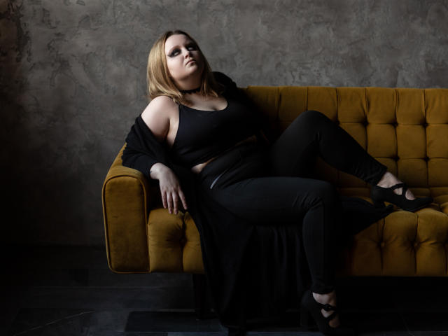 MissEmmar is a curvy woman with blonde hair, black top, and pants, sitting on a yellow couch, legs crossed, black shoes, and a black off-shoulder robe.