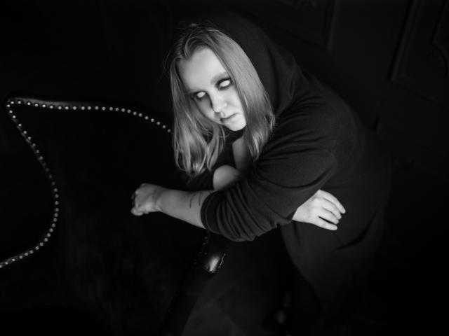 Black-and-white photo of MissEmmar with shoulder-length blonde hair, wearing a dark hoodie, arms crossed, sitting on a studded velvet chair, looking down with a serious expression.