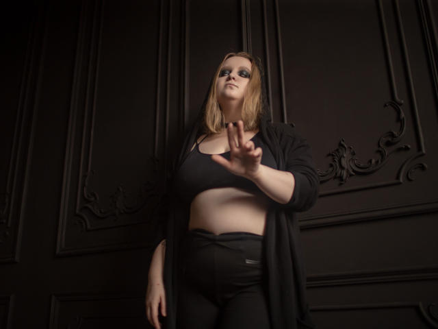 MissEmmar with blonde hair, black sports bra, and black pants, reaching out with a hand. She's wearing a black coat, and her tits are slightly visible under the bra.