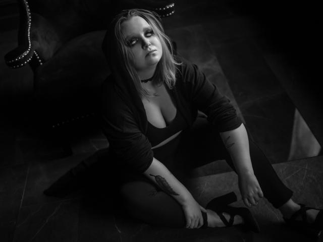 MissEmmar with pale skin, blonde hair, and tattoos, sitting on the floor in dark clothes, showing her big boobs and looking up with a serious expression. Black-and-white photo.