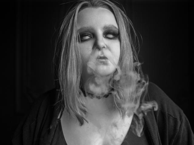 Black-and-white photo of MissEmmar with dirty blonde, messy hair, heavy eye makeup, and a choker, smoking with visible smoke in front of her face, wearing a dark shirt.
