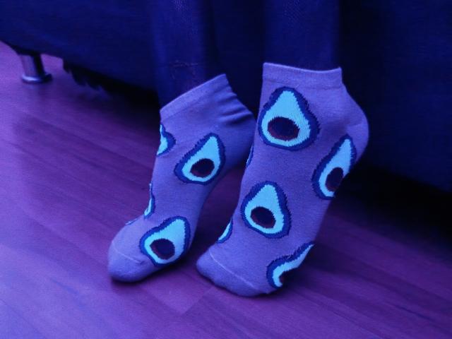 KiraFelot's feet in purple socks with blue and black peep-eye patterns, standing on pinkish wooden floor, under dark blue furniture.