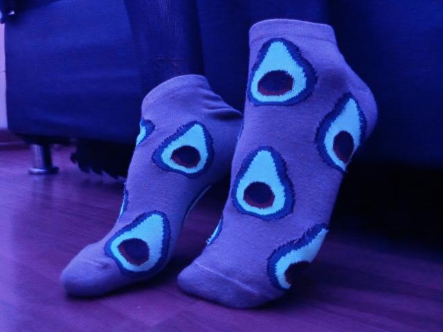 KiraFelot's feet are wearing gray socks with peacock eye patterns, standing on a wooden floor, under blue lighting. Socks are slightly stretched, showing their texture.