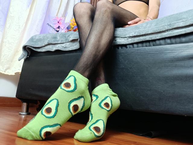 KiraFelot's legs in black fishnet stockings, wearing green socks with eye patterns, sitting on a bed with a grey cover. Wooden floor, partial bed visible.