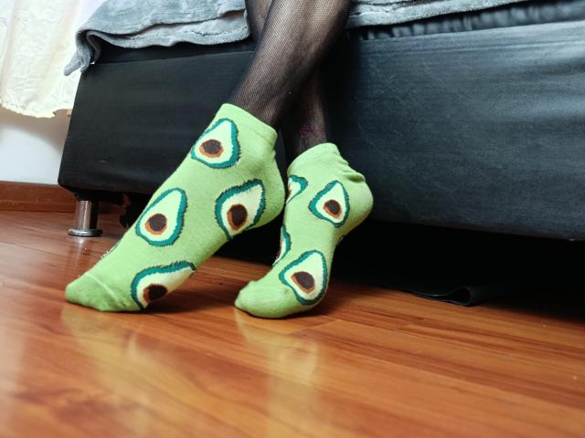 KiraFelot's feet in green socks with eye patterns, wearing black pantyhose, standing on wooden floor, legs against black leather couch, socks slightly wrinkled, wooden floor reflecting light.