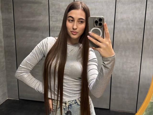 LateshaLuckenbaugh with long brown hair, wearing a grey long-sleeve top, holding a phone, taking a mirror selfie, hands on hips, in a grey-walled room.