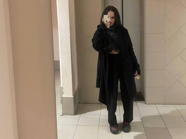 AlaneHarradon has dark hair, wearing a black coat, black top, black pants, and red slip-on shoes, holding a phone, standing in a tiled hallway.