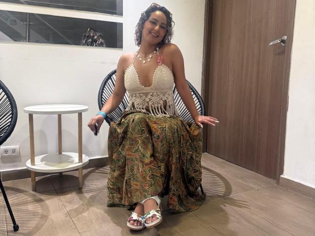 Flora Saenz mit lockigem, braunem Haar, trägt eine weiße Spitzenbluse, einen farbenfrohen, gemusterten Rock und weiße Sandalen. Sie sitzt auf einem schwarzen Stuhl, lächelt und hält eine Fernbedienung. Ihre Brüste sind durch die Spitze sichtbar..