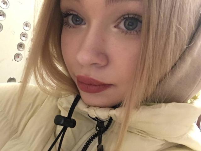 AnnamaeSvehla's face with blue eyes, red lipstick, and blonde hair, wearing a cream hoodie with a black strap, looking at the camera. Her tits are slightly visible.