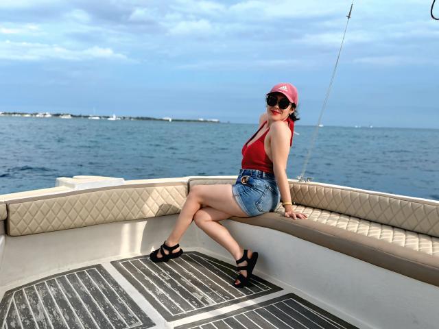 MabyLegrand, met een rood tanktop, spijkerbroekshorts, zwarte hakken en een rode pet, zit op de beklede rand van de boot, waarbij haar achterwerk zichtbaar is, met gekruiste benen, en draagt een zwarte zonnebril, met op de achtergrond de blauwe oceaan..