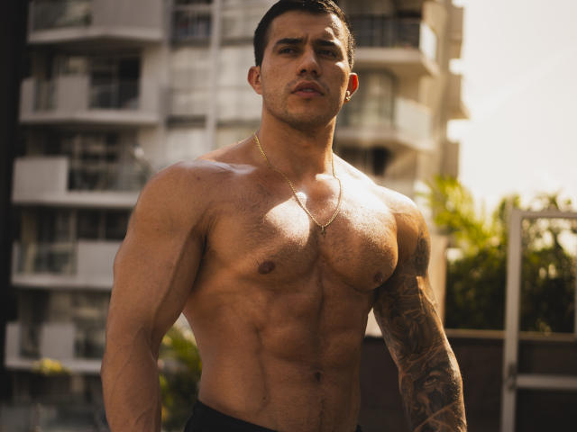 Muscular GarrettAlvin with a tattooed arm, short dark hair, and a gold necklace, standing shirtless, showing defined abs and pecs, wearing black pants, sunlight on his chest.