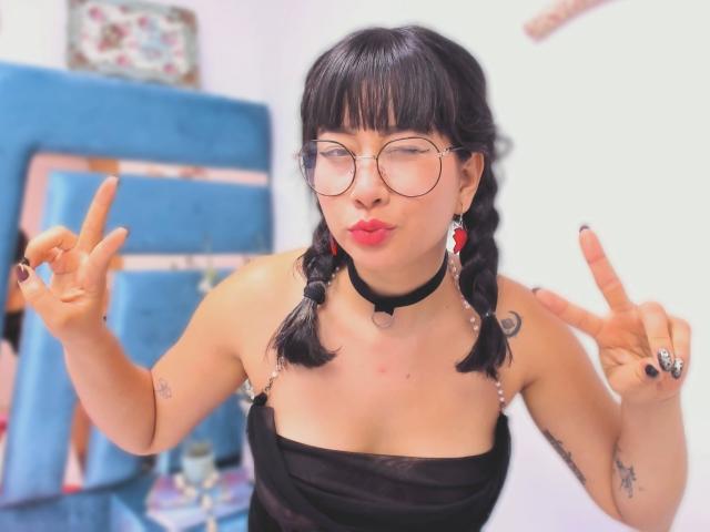 Genesiss, con coletas negras, lápiz labial rojo, gafas, collar negro y un top negro sin tirantes, haciendo el gesto de la "V" con ambas manos, mostrando sus tatuajes y esmalte de uñas negro..
