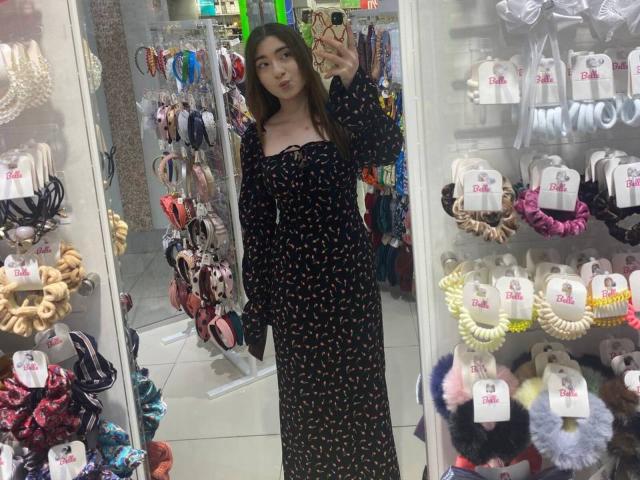 LiliaBilka, vestida con un vestido negro con pequeños lunares amarillos, sosteniendo un teléfono, está de pie en una tienda de joyería y accesorios, con pulseras de colores y collares blancos con cuentas..
