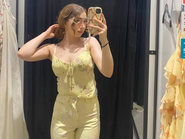 LiliaBilka mit Brille, trägt einen gelben, blumigen Overall, hält ihr Handy in einem Spiegel-Selfie, ihre Brüste sind leicht durch den Stoff sichtbar, ihr Hintern ist bedeckt..
