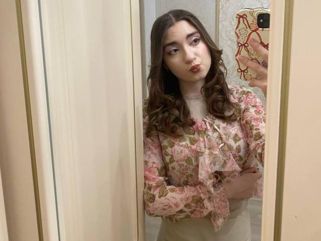 LiliaBilka met golvend bruin haar, een roze bloes met bloemenprint, volle lippen, die een foto van zichzelf in de spiegel maakt en een telefoon vasthoudt, waarbij haar borsten licht zichtbaar zijn onder de blouse, witte achtergrond, reflecterend spiegel..