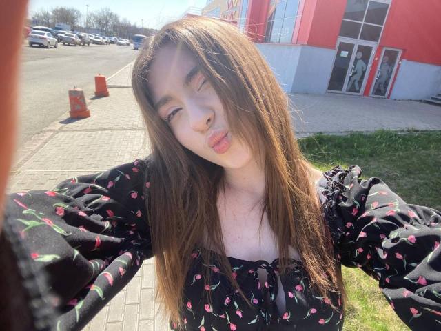 LiliaBilka met lang bruin haar, volle lippen en een zwarte jurk met bloemenprint, staat buiten, de zon schijnt, haar ogen zijn gesloten, ze heeft zwarte pofmouwen, een klein boezem, een schattig gezicht, en er is gras op de voorgrond..