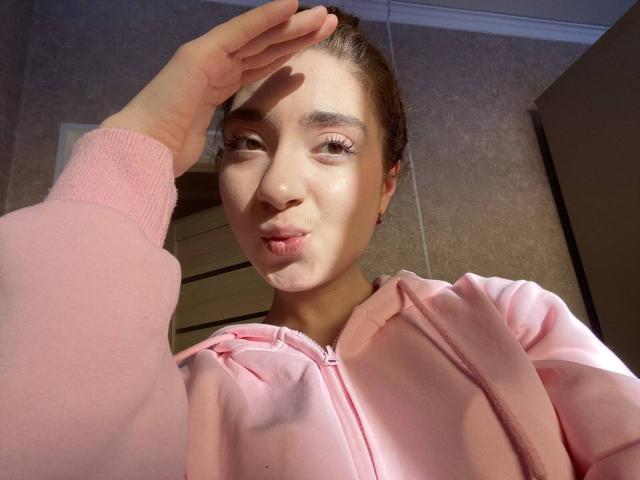 LiliaBilka, con una sudadera rosa hinchada, labios fruncidos, una mano en su cabello castaño, luz solar en su rostro, una nariz pequeña, piel clara y unos ojos rosados adorables, mirando a la cámara..